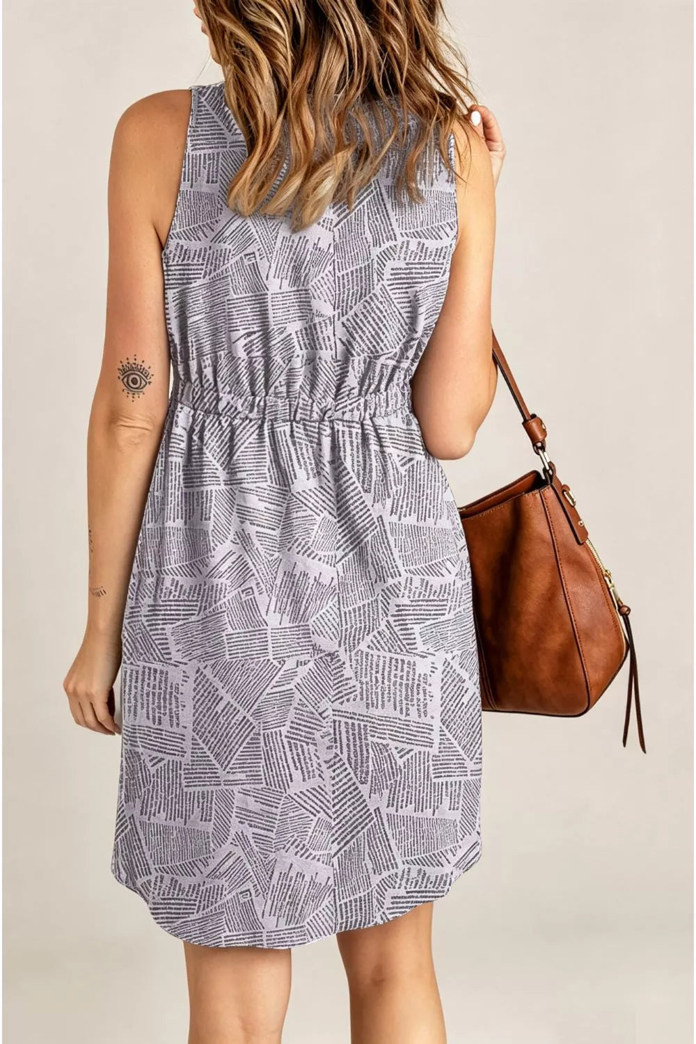 A woman shown from the back wearing a sleeveless grey and white abstract patterned mini dress with a gathered waist. She is carrying a brown leather shoulder bag against a plain beige studio background.