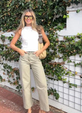 Model wearing wide leg beige coloured denim jeans with a white tank top and black sunglasses with a garden wall background