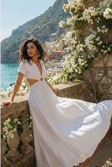 Woman in a white maxi dress standing on a scenic coastal path with flowers and mountains in the background