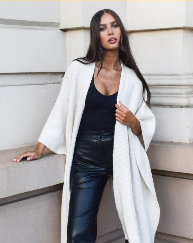 Woman wearing a white poncho cardigan over a black outfit, standing against a light-coloured wall.