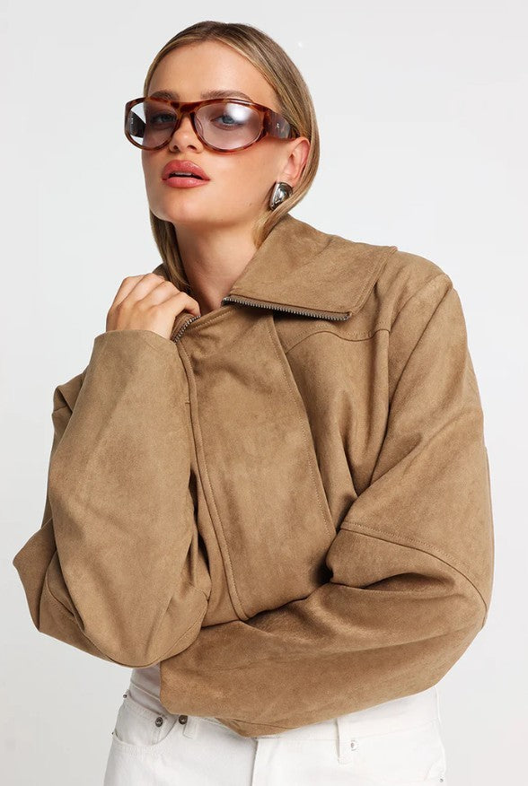 A woman wearing a cropped tan faux-suede jacket with a high collar and front zipper, styled with oversized sunglasses and white pants against a light gray studio background.