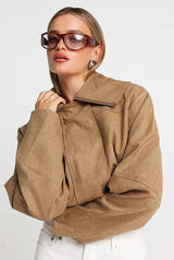 A woman wearing a cropped tan faux-suede jacket with a high collar and front zipper, styled with oversized sunglasses and white pants against a light gray studio background.