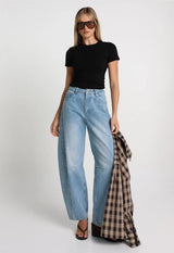 model with blonde hair wearing aviator style sunglasses wearing a black short sleeve top and light denim barrel jeans holding a checkered shirt