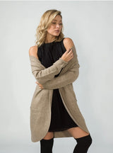 A blonde woman models a long, open-front ribbed knit wool blend cardigan in a light taupe-beige color, worn over a black off-the-shoulder dress and black knee-high socks, against a plain light grey studio background.
