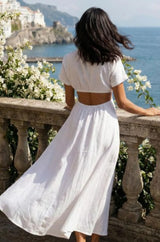 Woman in a white maxi outfit standing on a balcony overlooking a scenic coastal view with flowers in the foreground.