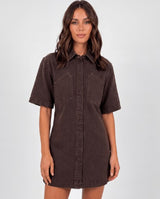 A woman modelling a dark brown short-sleeve button-up shirt mini dress with a washed denim-like texture, shown against a light grey studio background.