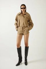 A model stands against a light gray studio background wearing a matching tan cropped jacket and shorts set, black knee-high boots and dark sunglasses.