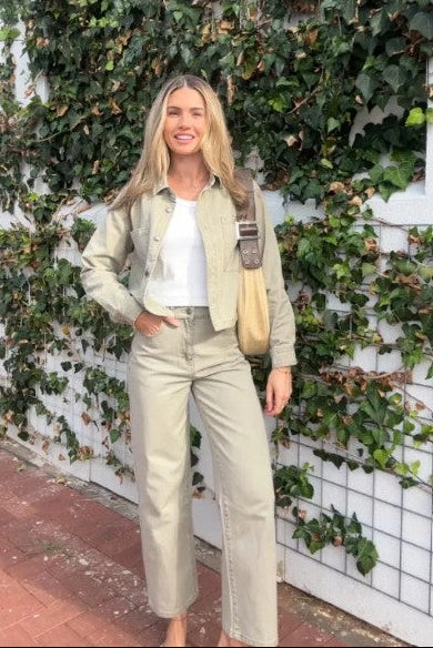 Model wearing wide leg beige coloured denim jeans with a white tank top holding a beige handbag with a garden wall background