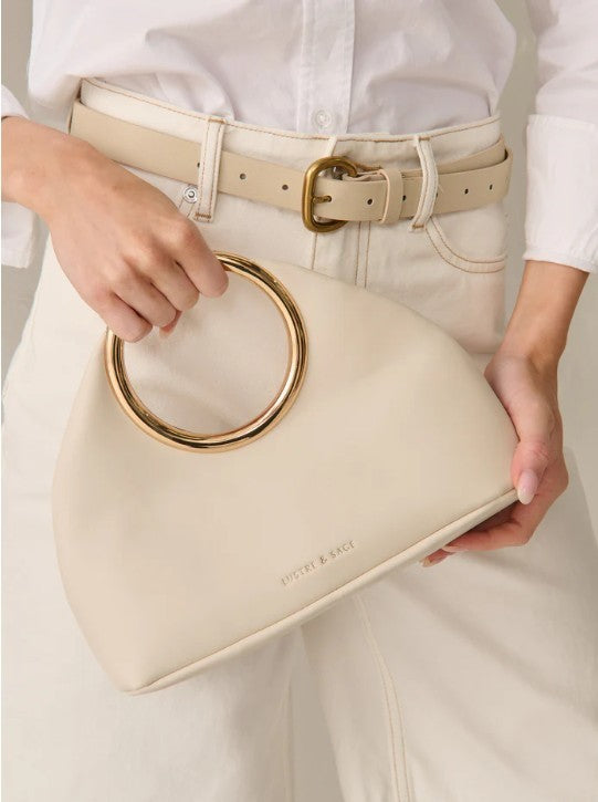 Beige handbag with gold ring handles held by a person wearing a white shirt and beige pants.