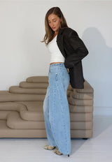 model with blonde hair wearing a crop white top, black jacket and light denim barrel leg jeans standing next to a light brown lounge on a white background.