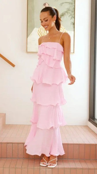 A woman wearing a light pink tiered ruffle maxi dress with spaghetti straps, standing on a tiled staircase indoors.