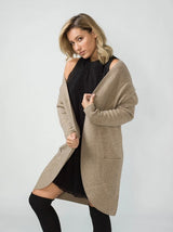 A woman modelling a long, open-front ribbed knit wool blend cardigan in a light taupe-beige color, worn over a black dress and black over-the-knee socks, against a plain light grey studio background.