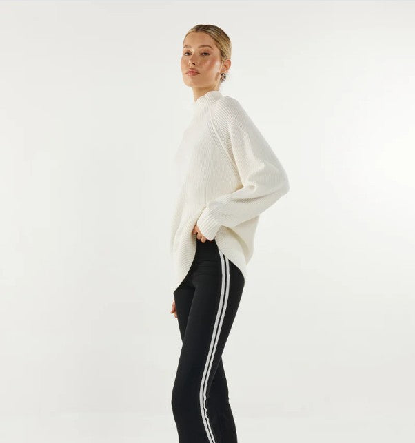 Woman wearing a white turtleneck sweater and black leggings with white stripes on a light grey background