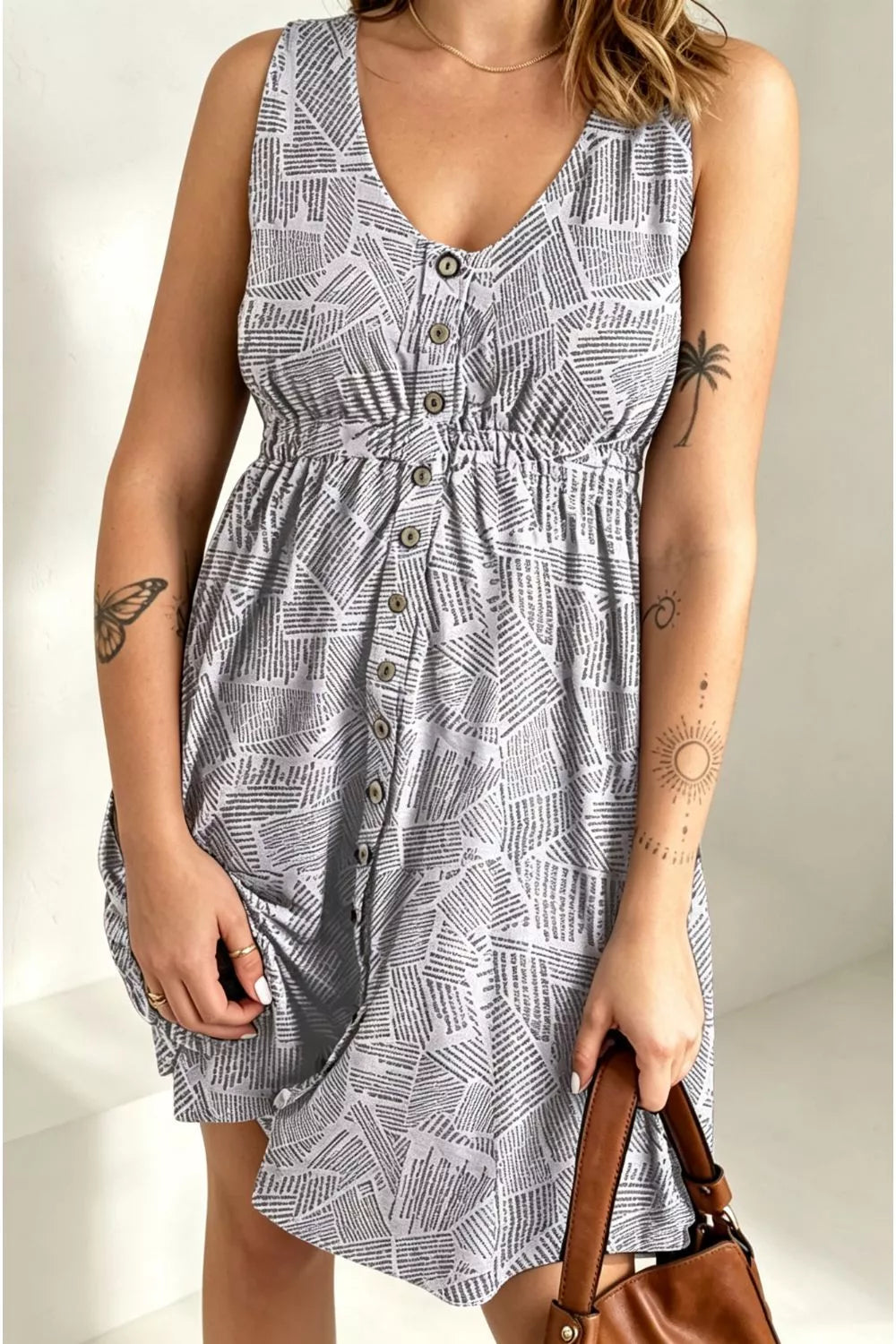 A sleeveless V-neck dress with a grey and white geometric line pattern, featuring a button-down front and a gathered waist, shown on a model holding a brown handbag.