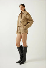 A model stands against a light gray studio background wearing a tan cropped jacket and matching shorts, paired with black knee-high heeled boots.