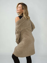A woman wearing a chunky, oversized taupe ribbed knit wool blend cardigan, photographed from a side-back angle against a light grey studio background.