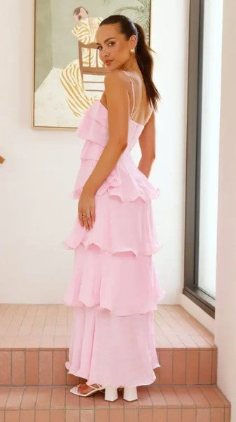 A woman wearing a light pink tiered maxi dress with spaghetti straps and a ruffled back, standing on terracotta steps in a bright indoor setting.