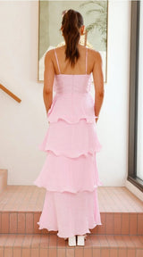 A woman viewed from behind wearing a light pink tiered maxi dress with thin spaghetti straps, standing on tiled steps in a bright indoor setting.