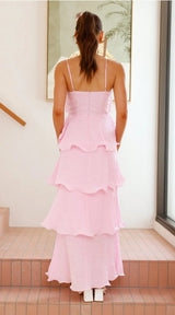 A woman viewed from behind wearing a light pink tiered maxi dress with thin spaghetti straps, standing on tiled steps in a bright indoor setting.
