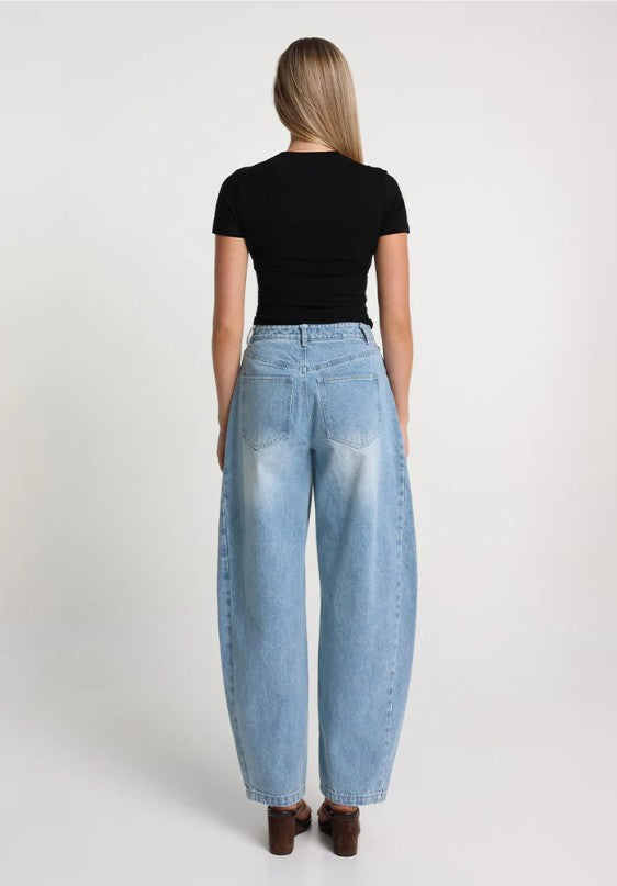 model with blonde hair wearing a black short sleeve top and light denim barrel jeans standing with her back to the screen on a white background