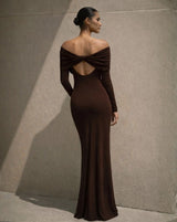 A woman modeling an off-the-shoulder, long-sleeved floor-length gown in a deep chocolate brown color, shown from the back against a textured concrete wall.