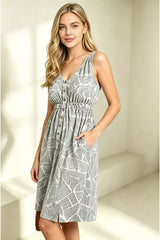 A woman modeling a sleeveless V-neck mini dress with a grey and white geometric line pattern, button front, and side pockets in a bright, minimalist room.