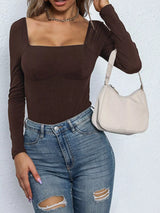 Person wearing a brown long-sleeve bodysuit and blue jeans, holding a beige handbag against a white background.