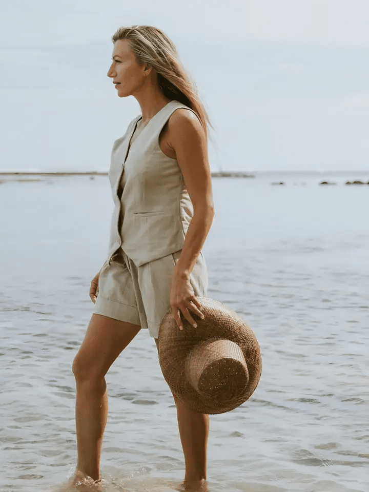 A model wearing the Carrie Sleeveless Vest in Natural Beige, walking along the shoreline with a straw hat in hand.