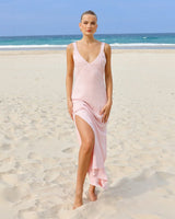 A woman stands on a sandy beach wearing a light pink sleeveless maxi dress with a V-neckline and a high side slit.
