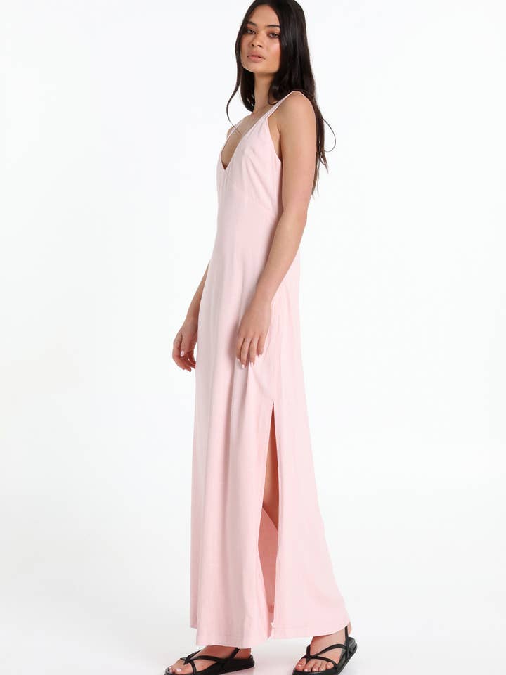 A woman modelling a light pink sleeveless maxi dress with a V-neckline and a high side slit, paired with black sandals, against a plain white studio background.