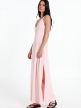 A woman modelling a light pink sleeveless maxi dress with a V-neckline and a high side slit, paired with black sandals, against a plain white studio background.