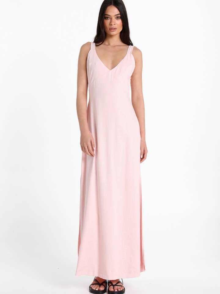 A woman modelling a light pink sleeveless maxi dress with a V-neckline and a high side slit, paired with black sandals, against a plain white studio background.