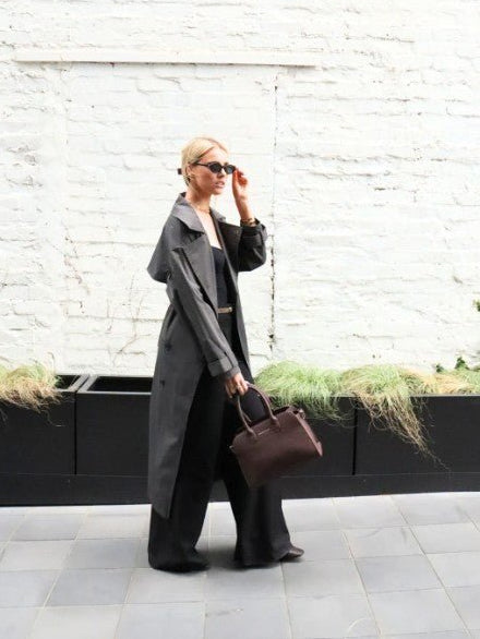 Person wearing a long coat and holding a brown bag against a white brick wall.
