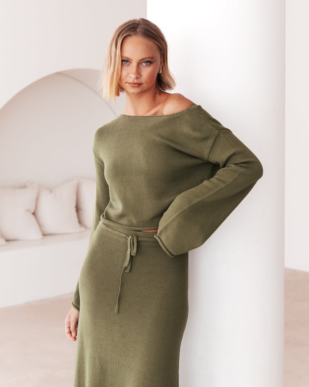 A model wearing the Cora Knit Set in olive green, leaning softly against a white wall in a relaxed pose.