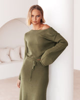 A model wearing the Cora Knit Set in olive green, leaning softly against a white wall in a relaxed pose.