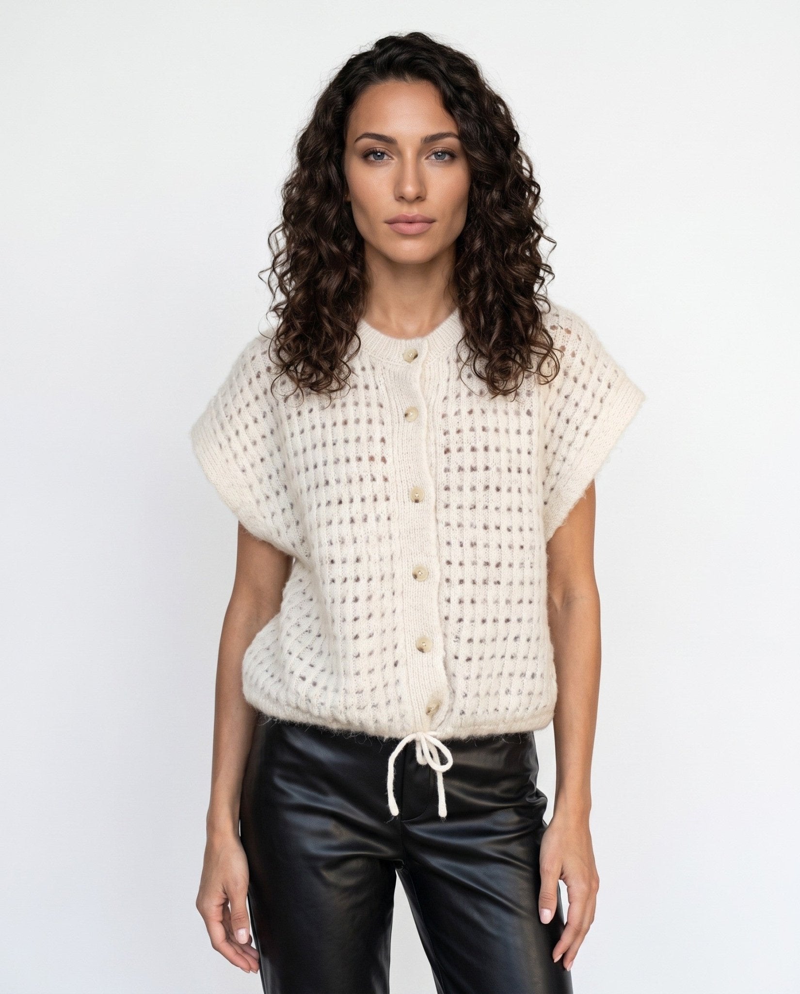 A woman with curly dark hair wearing a cream-colored, short-sleeved open-knit button-up cardigan and black leather pants, standing against a plain white studio background.