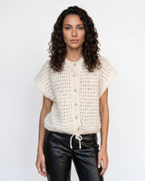 A woman with curly dark hair wearing a cream-colored, short-sleeved open-knit button-up cardigan and black leather pants, standing against a plain white studio background.