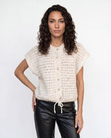 A woman with curly dark hair wearing a cream-colored, short-sleeved open-knit cardigan with a button-up front, paired with black faux-leather pants, against a plain white studio background.