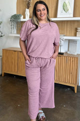 Woman wearing a pink loungewear pants and top set standing in a room with wooden furniture and decor.