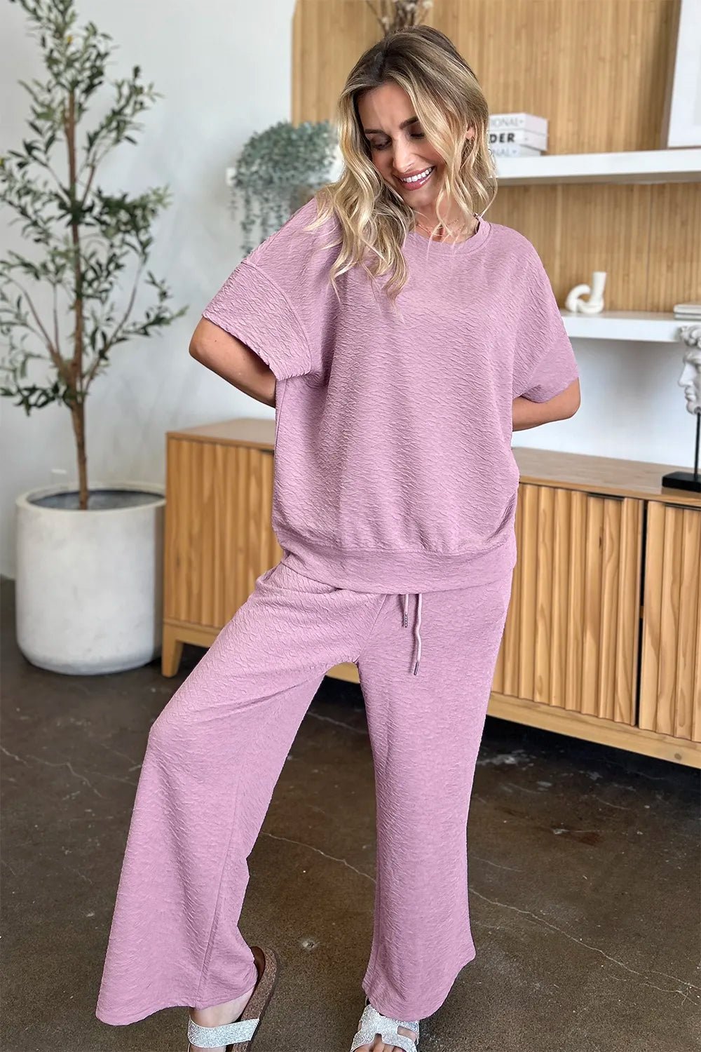 Woman wearing a pink loungewear pants and top set in a room with wooden furniture and a plant.