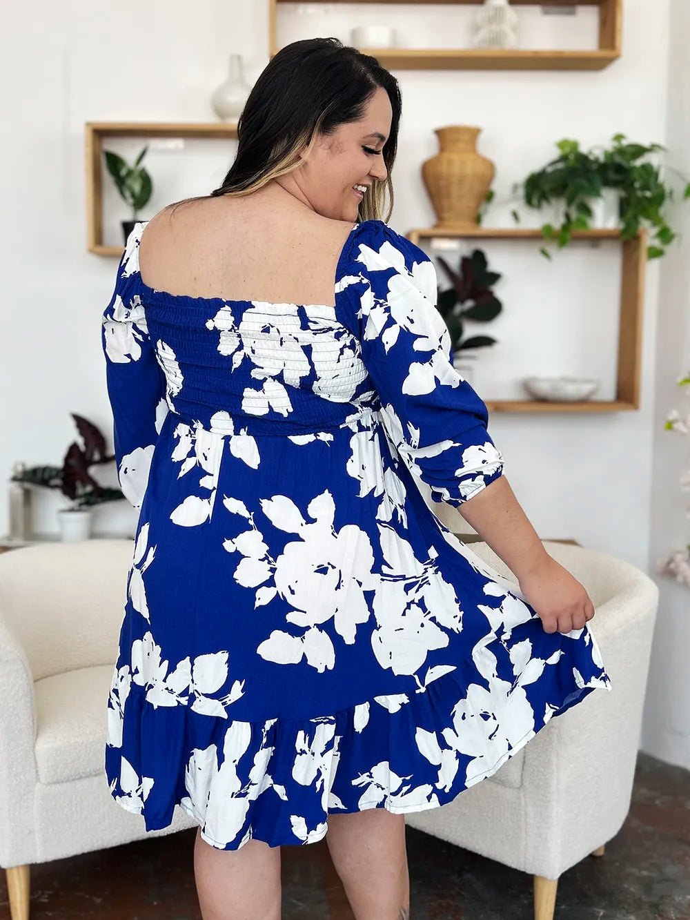 Woman wearing a blue floral dress sitting on a white chair in a room with plants and shelves.