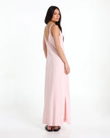 A woman modelling a light pink sleeveless maxi dress with a side slit, shown from a three-quarters back view against a plain white studio background.