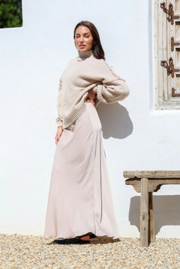 Woman wearing the Janelle Satin Maxi Skirt with matching knit jumper, standing against a white wall in sunlight.