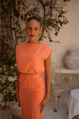 Woman in an orange orange top and skirt standing in a decorated indoor setting with plants and a chair.
