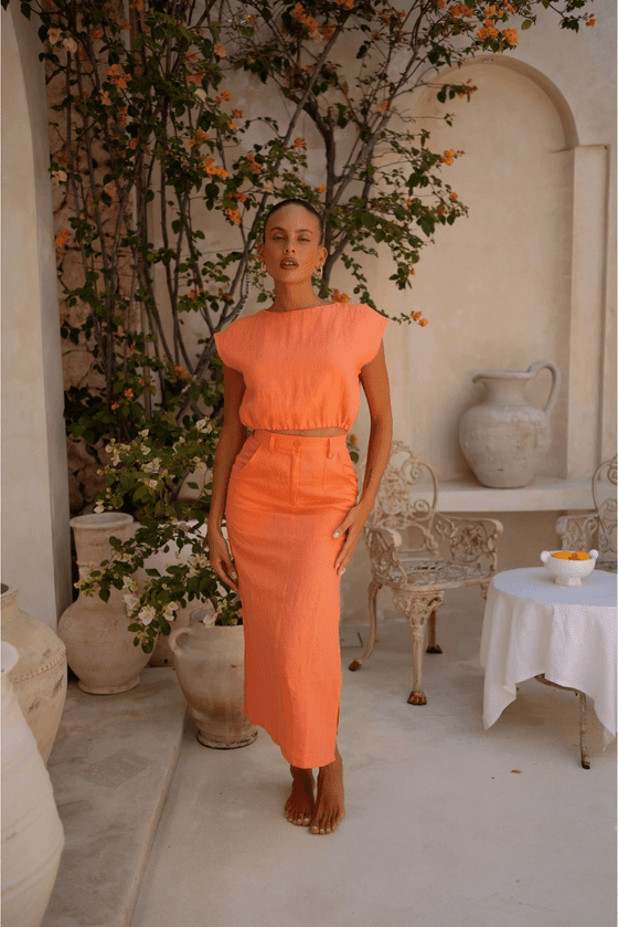 Woman in an orange orange top and skirt standing in a decorative outdoor setting with plants and a table.