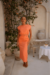 Woman in an orange orange top and skirt standing in a decorative outdoor setting with plants and a table.