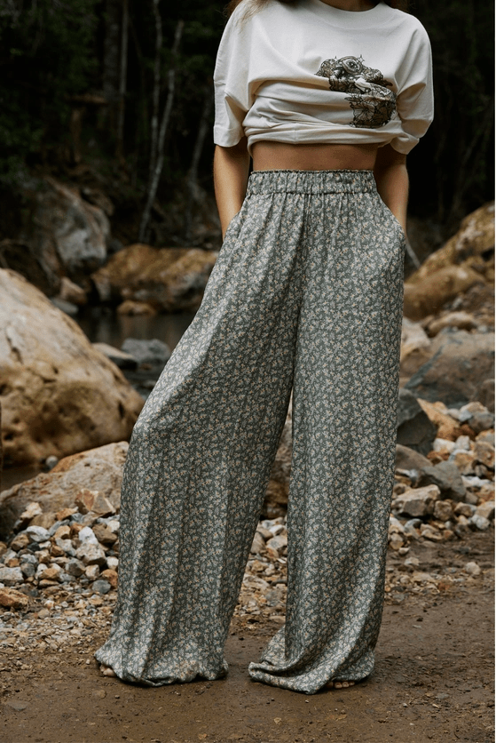 Person wearing a white crop top and patterned wide-leg pants standing in a natural setting with rocks and water.