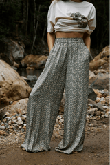 Person wearing a white crop top and patterned wide-leg pants standing in a natural setting with rocks and water.