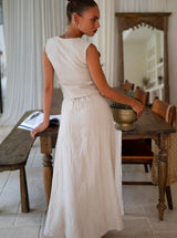 Woman in a beige sleeveless top and beige maxi skirt standing in a room with wooden furniture and a mirror.