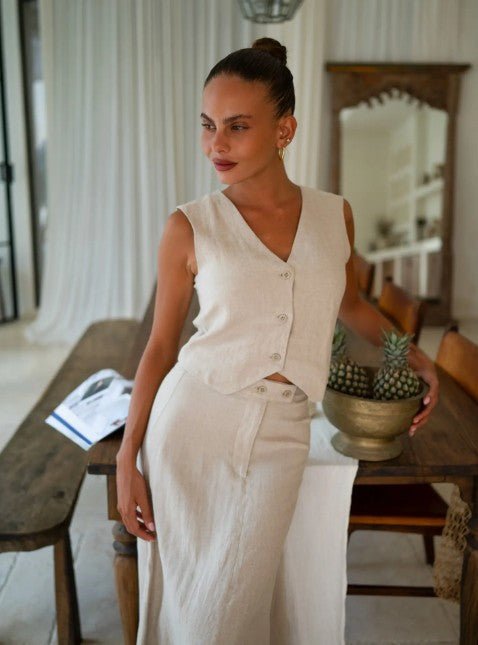 Woman in a beige sleeveless top and beige maxi skirt standing in a room with wooden furniture and decor.